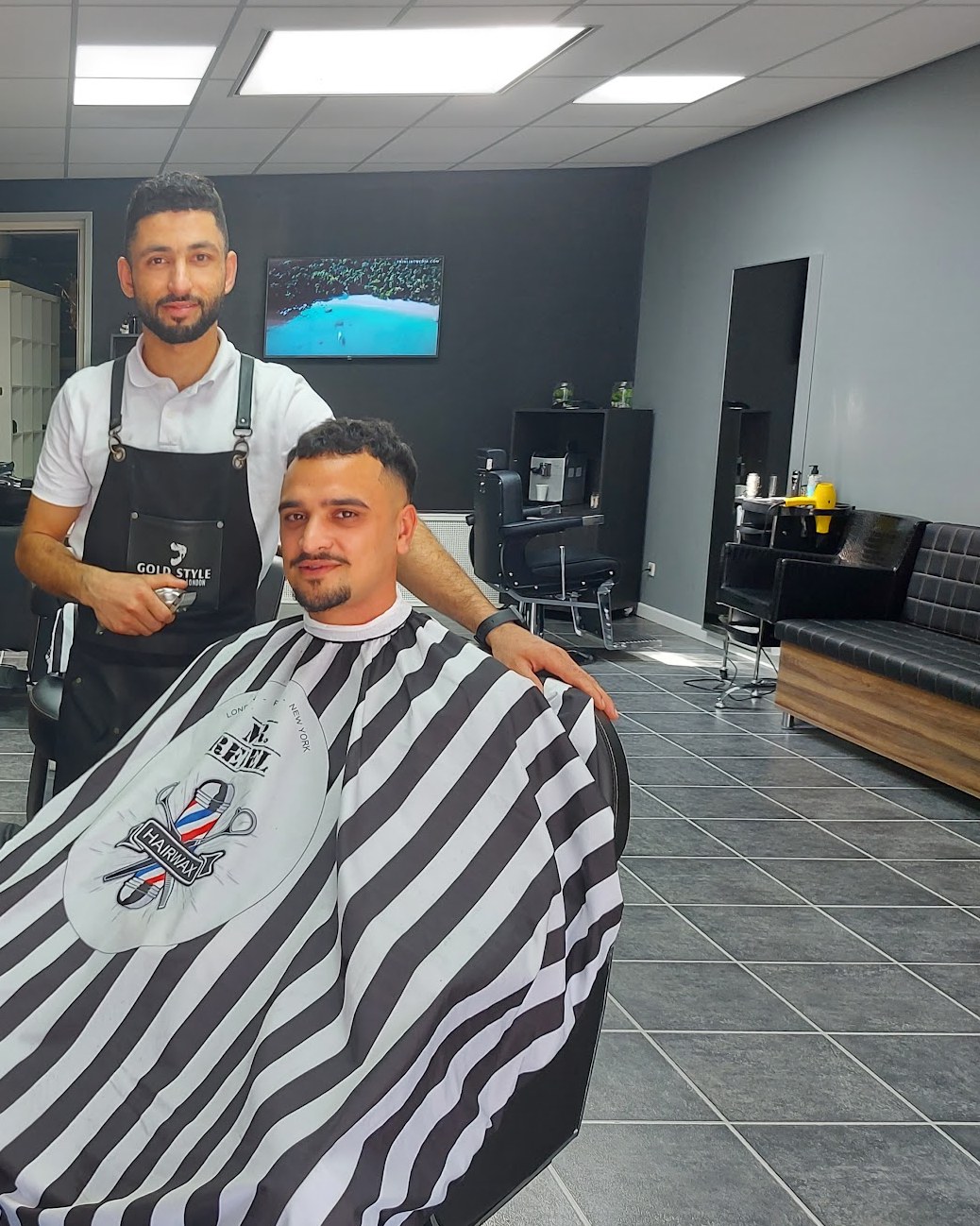 Een kapper van Kapsalon Hamoud achter een klant in een zwart-wit gestreepte kapmantel, in de zaak met donkere wanden en een tegelvloer